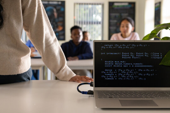 Indian instructor in cream knit teaching teens in class at cabled laptop showing code, copy space
