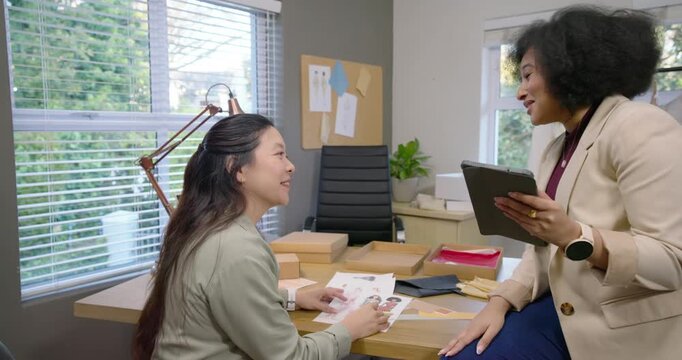 Diverse women team sharing tablet and comparing print mockups for design at desk