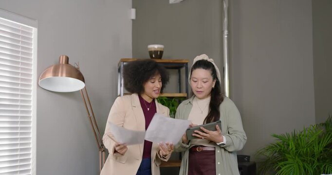 Diverse female team in officewear receiving papers, reviewing tablet, aligning notes at office