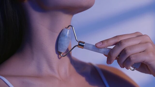 Placing jade roller below jawline, Asian woman massaging neck for skincare in room, thin strap top