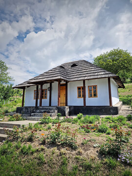 Traditional Moldovan peasant house with burned shingle roof. The cottage features white clay-plastered walls, wooden windows, and a porch supported by dark timber pillars, fronted by a blooming garden