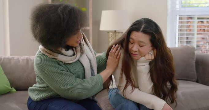 African American woman comforting Asian youth covering face, soothing shoulder on couch at home