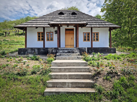 Traditional Moldovan peasant house with burned shingle roof. The cottage features white clay-plastered walls, wooden windows, and a porch supported by dark timber pillars, fronted by a blooming garden