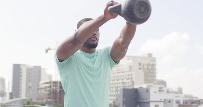 Mid-adult African man starting seated on rooftop railing, swinging kettlebell in reps for strength