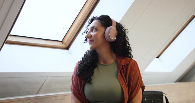 Woman following skylight, closing eyes, listening on pink headphones, scrolling phone near suitcase