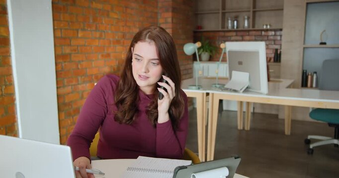 Female worker receiving phone call, taking notes, checking tablet, typing on laptop in office