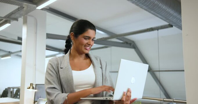 Adult Indian woman standing on mezzanine walkway typing on silver laptop while responding to camera