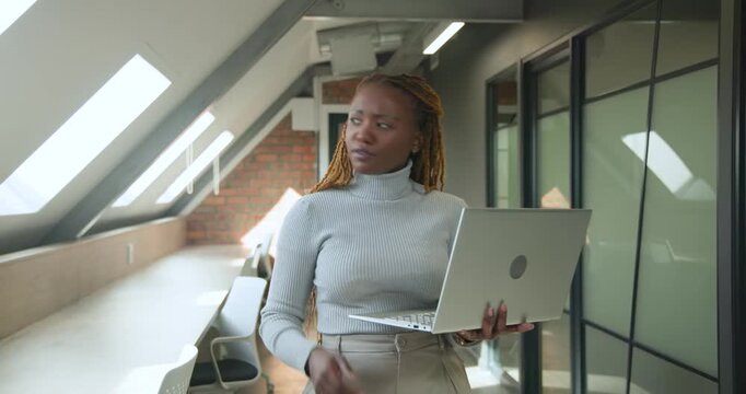 Mid-adult woman wearing grey turtleneck holding laptop in loft corridor, reading message, laughing