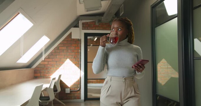 African woman in gray sweater entering, walking in loft holding pink phone and mug, reading message