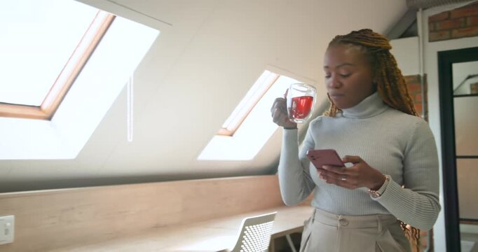 African woman standing in loft in turtleneck reading smartphone smiling and sipping tea, copy space