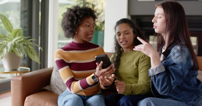 Diverse female friends leaning as left woman showing black phone, passing it for selfie on couch