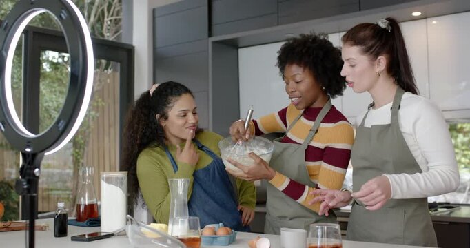 Diverse women whisking batter at kitchen island while ring light and DSLR entering frame, filming