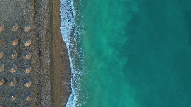 Aerial view of rows of beach umbrellas on a pebble shore with clear turquoise sea water in Greece.