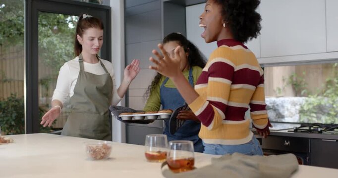 Diverse female friends timer beeping opening oven lifting tray with mitts celebrating bakes at home