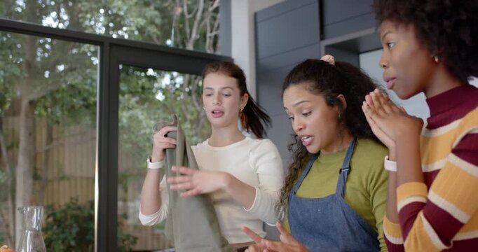 Three female friends unfolding green apron in kitchen, pointing at pastries and preparing to serve