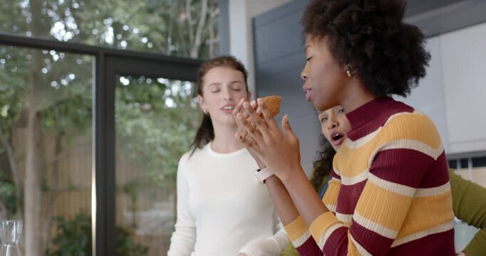 Three women pals watching woman in striped top picking muffin testing recipe in kitchen, copy space