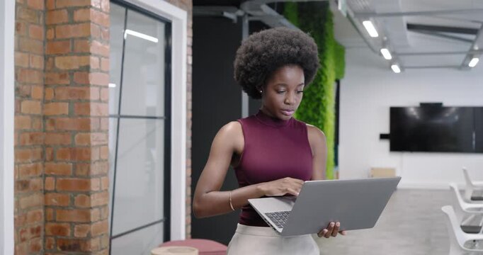 African American woman holding laptop, typing and adjusting laptop while working in open office