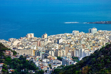 Naklejka premium Aerial view of Copacabana in Rio de Janeiro, Brazil, showing dense city buildings nestled between lush green mountains and the bright blue waters of the Atlantic Ocean.
