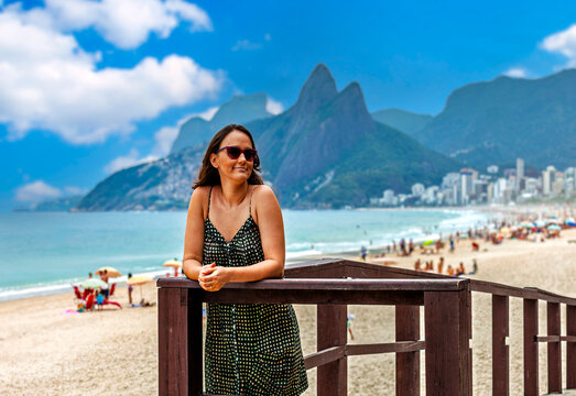 A happy woman with sunglasses leans on a railing overlooking the vibrant Ipanema beach in Rio de Janeiro, Brazil. The iconic Morro Dois Irm&atilde;os mountains dominate the scenic background.