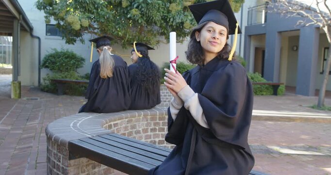 3 female graduates in gowns on courtyard planter main graduate lifting diploma turning for portrait