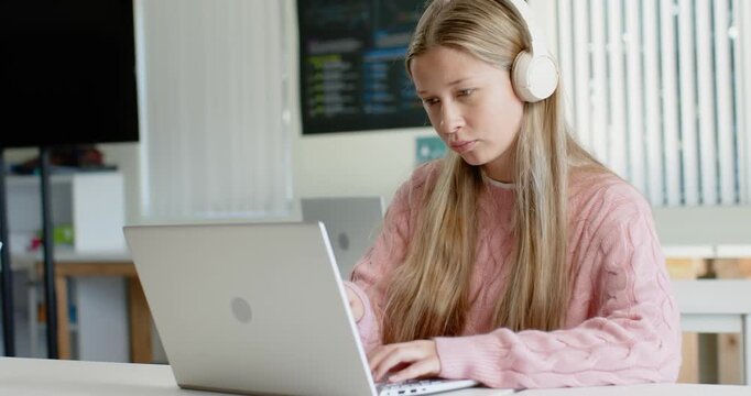 Teen female in pink knit with white headset reading laptop alert, pausing, typing at study table
