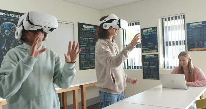 Diverse teenage female students putting on white VR headsets, reaching in lab, learning with laptop