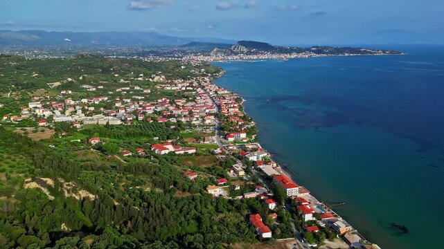 Aerial view of a coastal town with red-roofed houses nestled among lush green hills along the clear blue Mediterranean sea in Greece.