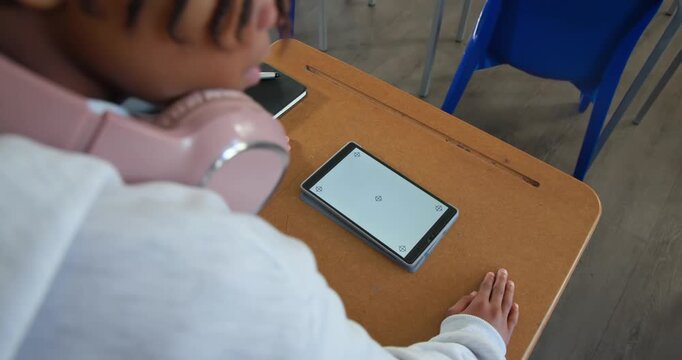 African American teenager adjusting pink headphones, leaning in classroom, checking tablet markers