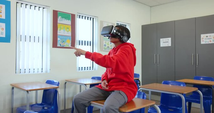 Headset on, African American teen in red hoodie sitting on desk in school gesturing, exploring VR