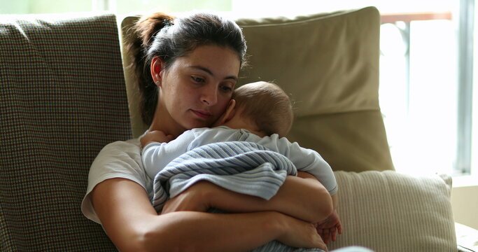Tired mother holding and caring for baby infant on couch. Exhausted mom falling asleep, candid and authentic