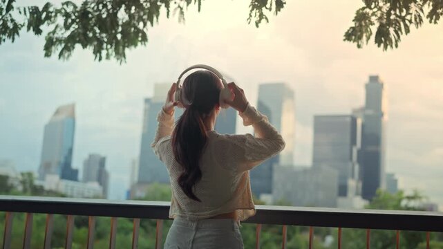 Back view of Asian woman putting on headphones dancing to music at city park balcony, cheerful girl enjoying wireless audio app with city building view at sunset for urban lifestyle mental wellness
