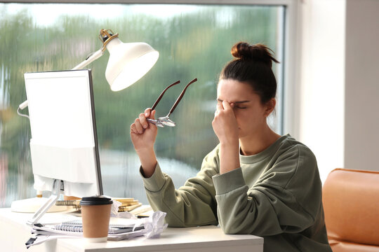 Young woman experiencing eye strain and headache from prolonged computer work at a messy desk, holding her glasses, Challenges of remote work and digital screen time