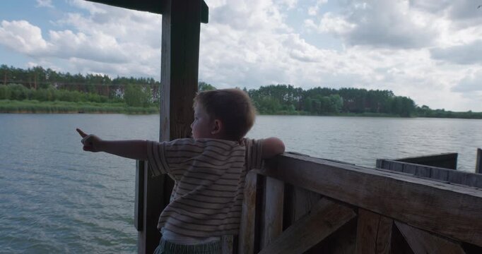 Cute little boy standing on wooden pier and pointing finger towards lake