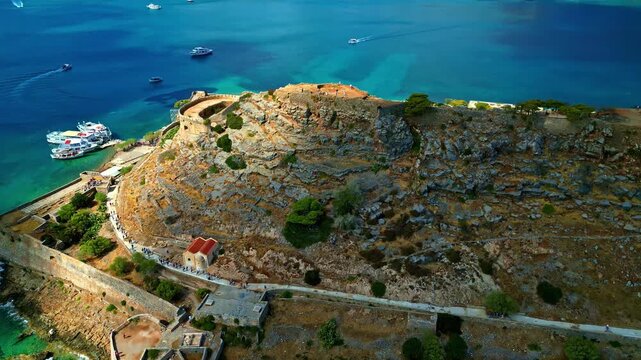 Aerial view of the historic Spinalonga fortress on a rocky island surrounded by turquoise sea with boats docked at the pier in Greece.