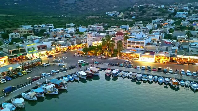 Aerial view of boats moored at the harbor quay with illuminated waterfront shops and restaurants in Crete, Greece.