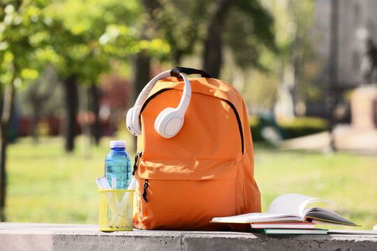 Orange backpack with headphones, school stationery and water bottle on ledge in park