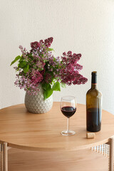Obraz premium Red wine bottle and glass with a bouquet of fresh purple lilac flowers in a vase on a wooden table, the arrangement sits on a round wooden table, with a cork resting beside the bottle
