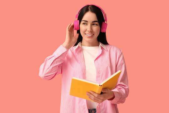 Young woman in headphones listening to audiobook on pink background