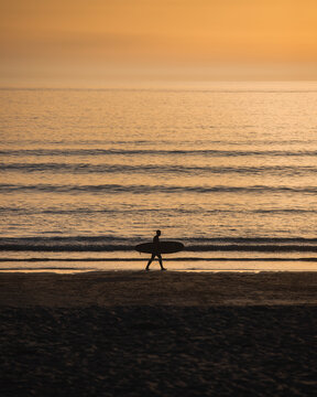 Penzance, England, United Kingdom - 18 July 2021: View of a surfer in silhouette carrying a surfboard along the sandy shore of Gwenver Beach during a golden sunset.