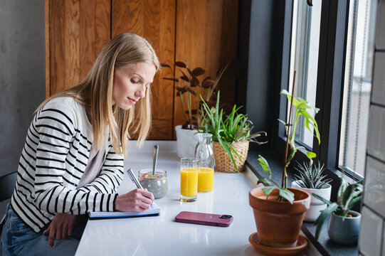 Young woman writing in a notebook while enjoying a healthy breakfast with chia pudding and fresh orange juice by the window in a cozy modern kitchen, symbolizing planning and mindful living