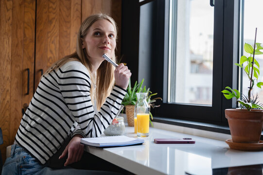Thoughtful young woman taking a break with a healthy breakfast while planning ideas by the window. Modern home office, productivity, wellness and creative lifestyle concept