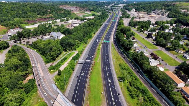 Aerial view of highway traffic moving through suburban hills with roads, homes, trees, and industrial areas, representing transportation infrastructure and connectivity.