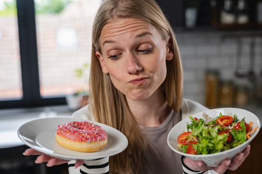 Confused woman deciding between a sugary donut and a healthy salad at home. Concept of diet, nutrition, cravings, self-control and balanced lifestyle