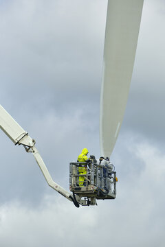 Esbjerg, Syddanmark, Denmark - 24 July 2026: View of crew performing maintenance on a wind turbine blade using a high-reaching cherry picker against a cloudy sky.