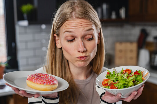 Young woman comparing a pink frosted donut and fresh salad in a modern kitchen. Healthy choices, balanced diet, temptation, nutrition and wellness lifestyle concept