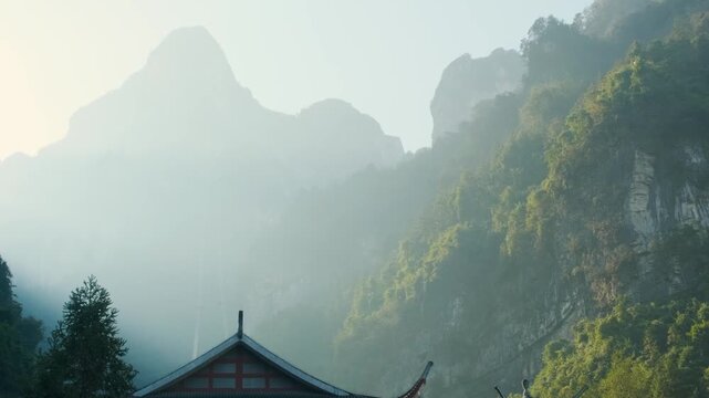 Traditional Chinese architecture stands beneath towering peaks of Tianmen Mountain in Zhangjiajie, Hunan, China with misty cliffs, forested mountains at sunrise. Travel destination in Asia