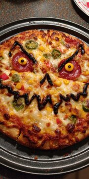 Spooky Halloween pizza decorated with a scary monster face using pepperoni and balsamic glaze on a round metal baking tray for festive party food