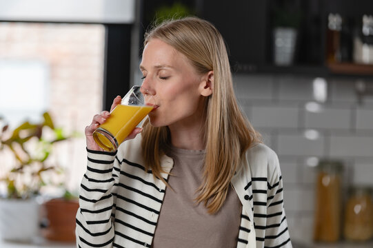 Young woman drinking fresh orange juice in a modern kitchen. Healthy lifestyle, nutrition, morning routine and wellness concept with natural light