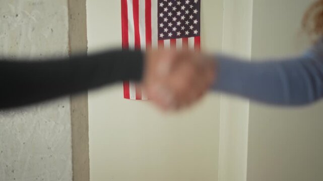 Two women shaking hands, hands visible and clasped at midframe, one wearing a black sleeve and one wearing a blue sleeve, american flag on the wall in a building; solidarity trust.