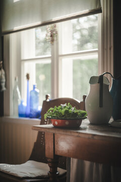 Laane County, Estonia - 02 June 2024: View of a red polka-dot bowl with fresh greens and a white pitcher on a rustic wooden table by a sunlit window with blue glass bottles.